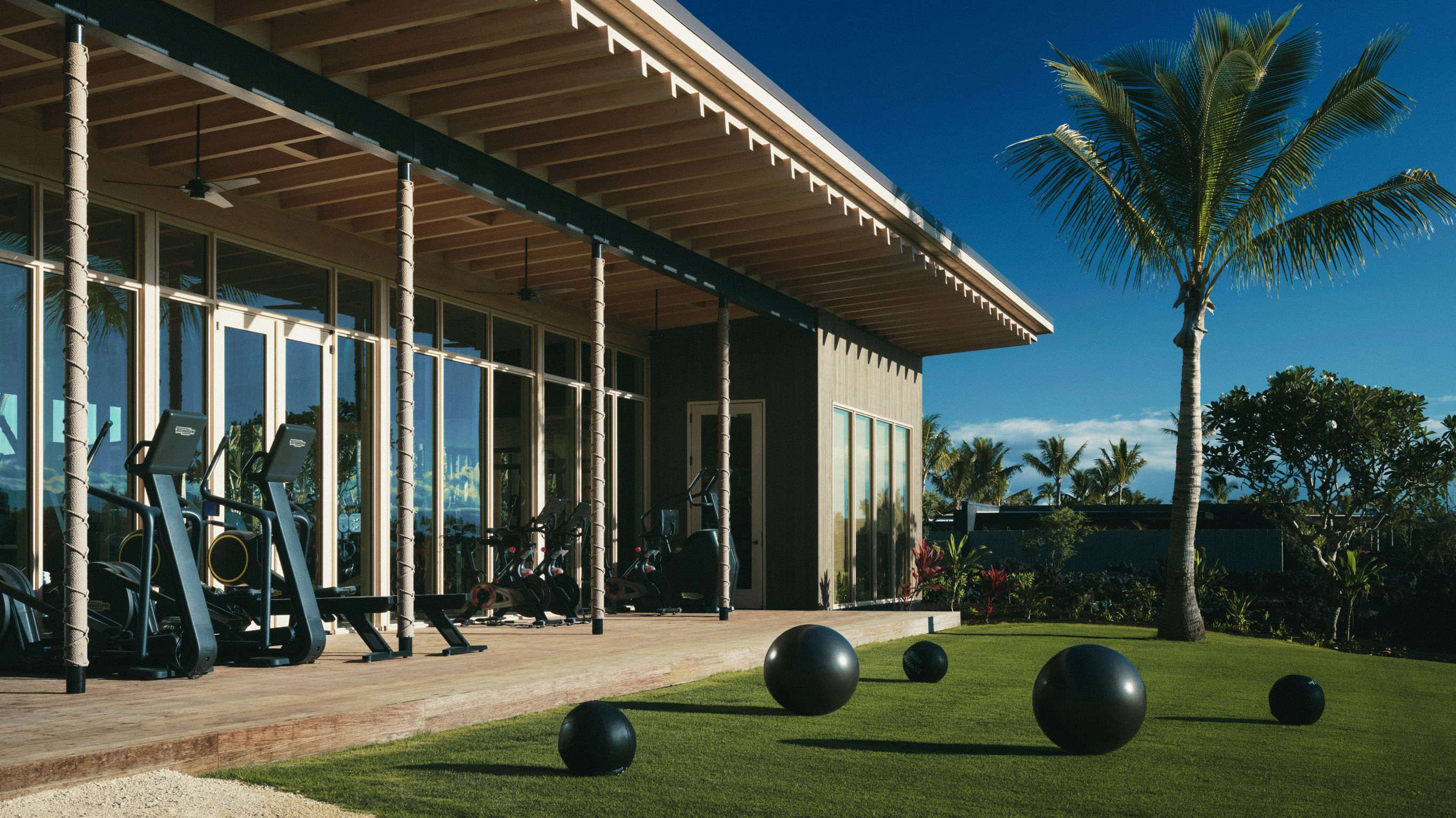 Open-air gym with cardio equipment facing palm trees under bright Hawaiian sunshine.